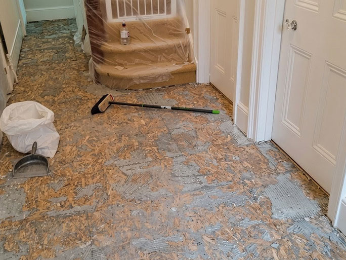 A hallway floor undergoing renovation, showing old plywood underlayment and adhesive residue being removed to expose original pine floorboards beneath