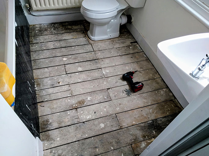 An old, weathered bathroom pine floor before restoration, showing stained, graying floorboards