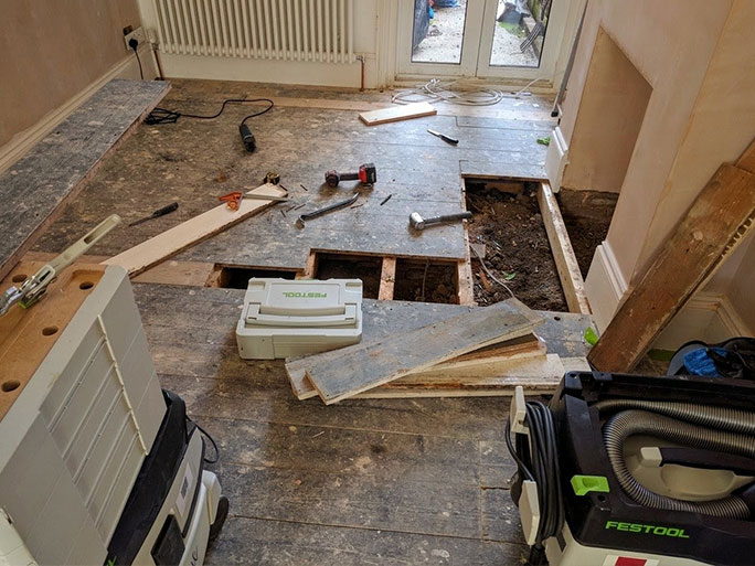 Damaged pine floorboards being removed during a floor restoration project to be replaced with reclaimed matching planks while exposing the underlying joists and dirt subfloor