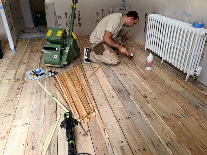 A flooring professional filling gaps in old pine floorboards by wedging wooden pine slivers between the planks in preparation for sanding and oil finishing the floor