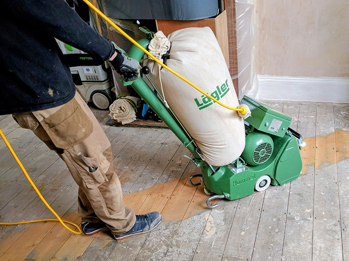 Using a Lägler Hummel belt sander the wood floor specialist starts sanding the pine floorboards to remove old dirt and finish, revealing the clean, light wood surface beneath